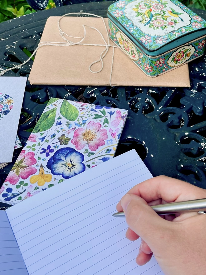 Hand with Pen Leaning on Botanical Inspired Notebook, Revealing Lined Pages, Smaller Notebook Nearby, on Garden Table