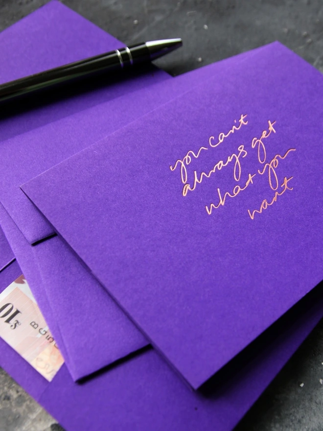 Hand foiled cash card or money wallet in a royal purple colour handfoiled in shiny rose gold foil text which says ‘You Can’t Always Get What You Want’.