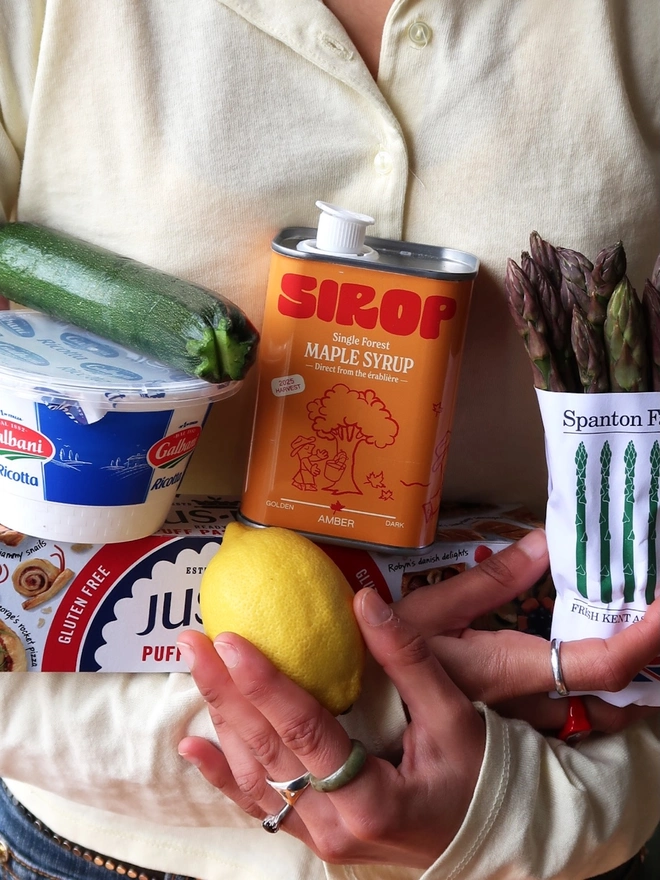 woman holding array of food items, including a metal container with orange label, which reads 'sirop single forest maple syrup'