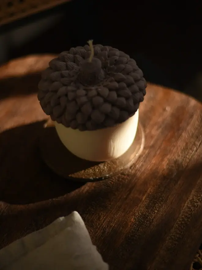 The Acorn Candle. A top-down view of an acorn candle with a chocolate brown textured cap, sitting on a wooden tray in warm, low sunlight. The woodland-inspired design brings rustic charm to a coffee table or side table.