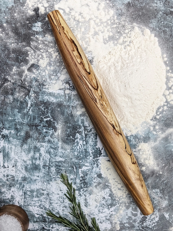 A stunning hand made rolling pin in Spalted Beech by Something From The Turnery. The rolling pin sits on an art house kitchen top amongst flour, rosemary and a wooden salt pot, prepped to bake some delicious bread! Made from Spalted Beech, the rolling pin features tapered ends amongst it’s own , never repeated natural grain patterns, travelling through golden browns, tans and stunning black lines to outline the changes in the timber. 