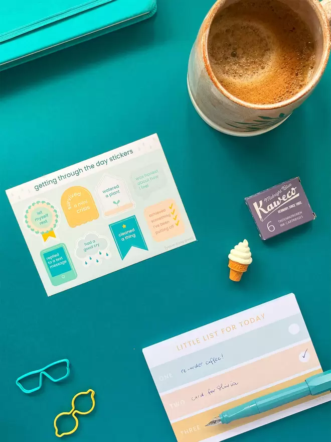 A colourful A6 sticker sheet sits on a blue background,surrounded by stationery items and a cup of coffee