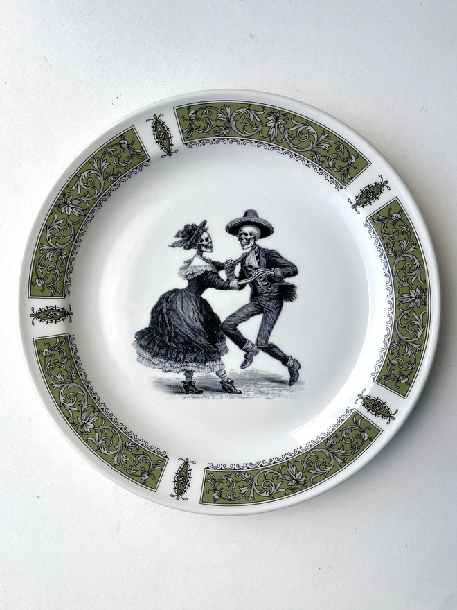 vintage plate with an ornate border, with a printed vintage illustration of two dancing day of the dead skeletons wearing a dress and a suit in the middle