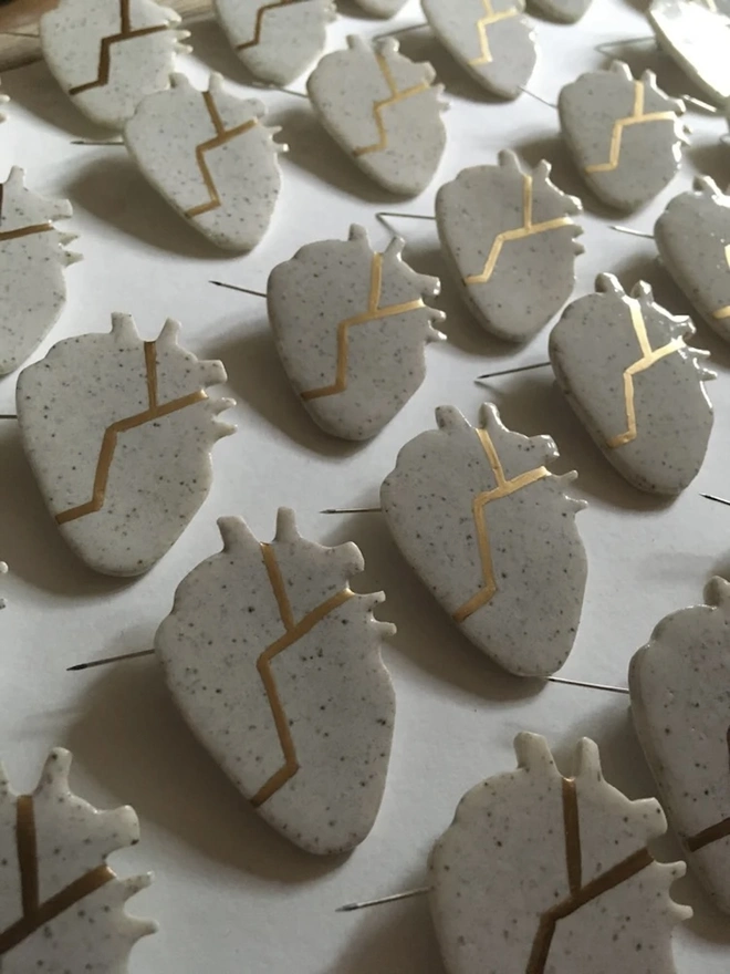 (Un)Broken Heart Pin, a group of white and gold heart pins sitting on a white surface. 