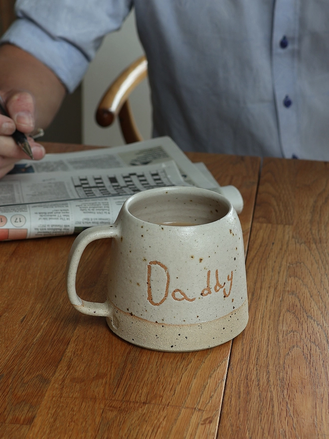 Personalised Kids' Handwriting Handmade Stoneware Mug with the name 'Daddy' on a wooden table with a man sat behind