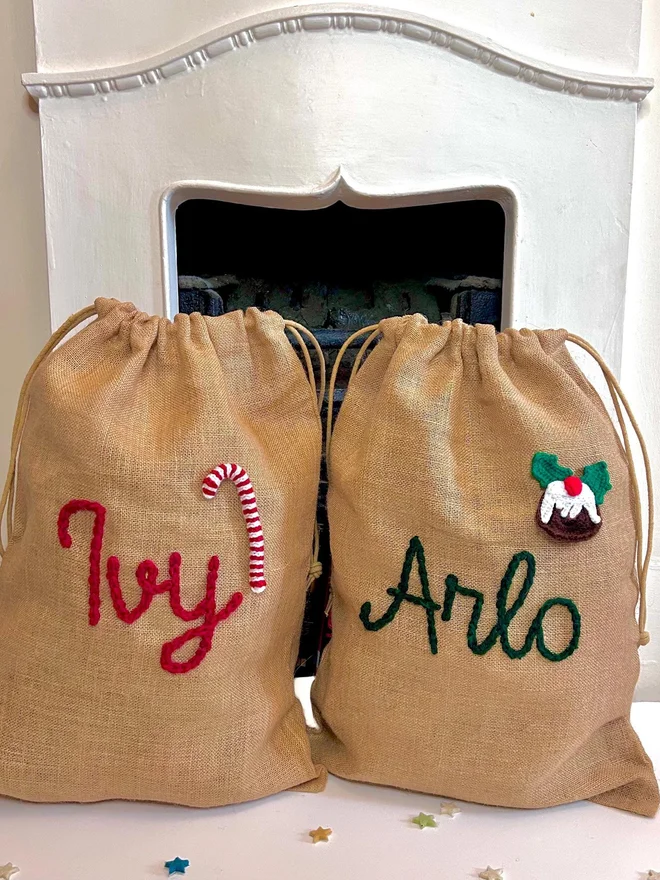 Personalised Christmas Sacks Landscape, two Christmas sacks sitting by a fireplace on a decorated floor. Each has their own personalisation. 