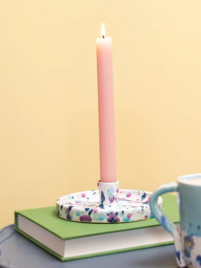Sweetpea Candle Stick Holder, a colourful candle holder with a pink lit candle in it on a book.It sits against a yellow backdrop. 