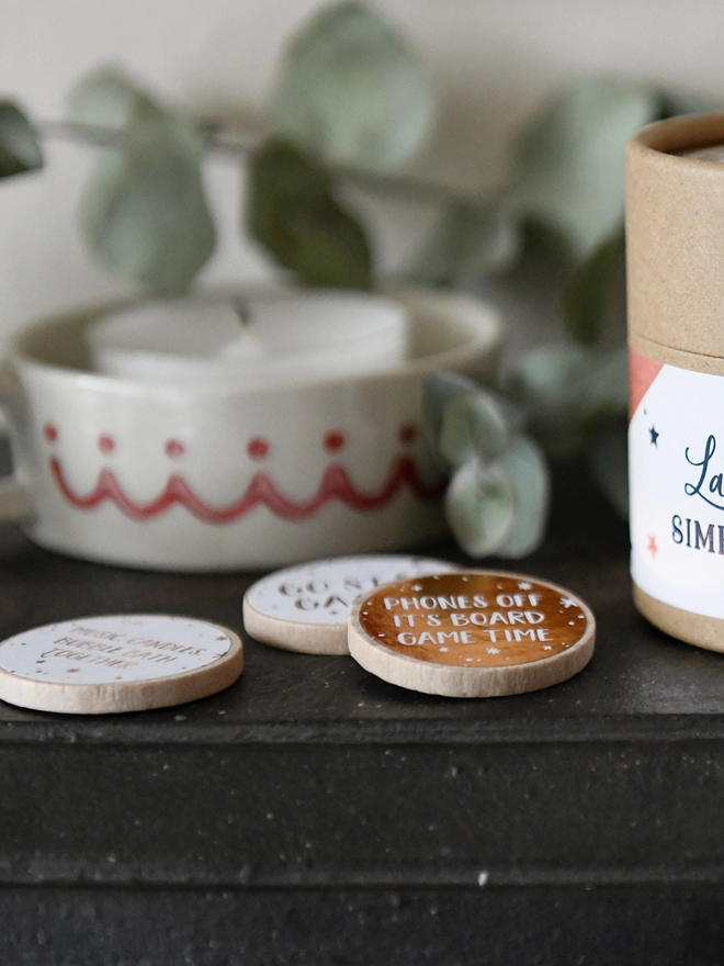 Several wooden tokens with copper foiled labels rest on a black mantlepiece beside a luxury cardboard jar.