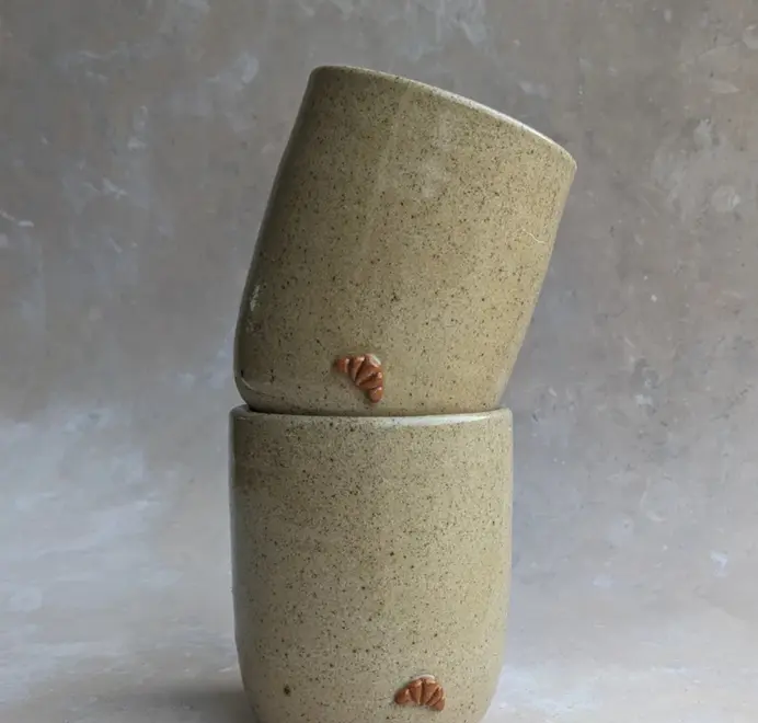 Two biscuit coloured croissant coffee cups with a subtle croissant design, placed against a grey marble background.