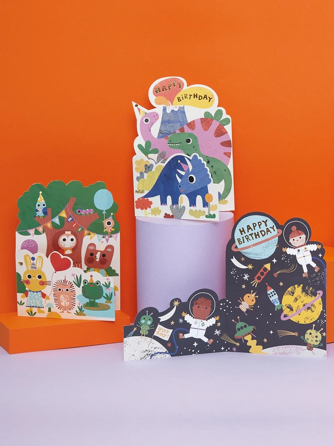 Other cards from the Raspberry Blossom ‘Fantabulous’ children’s greeting card collection. Grouped together on lilac plinths and with an orange background, the colourful birthday cards include a fun dinosaur, forest tea party, and space themes