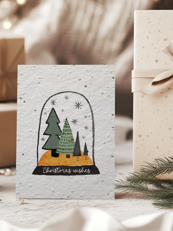 Christmas card with an illustrated snow globe on it with trees, snowflakes and 'Christmas Wishes'. The card is surrounded by presents, a pine cone and pine leaves.