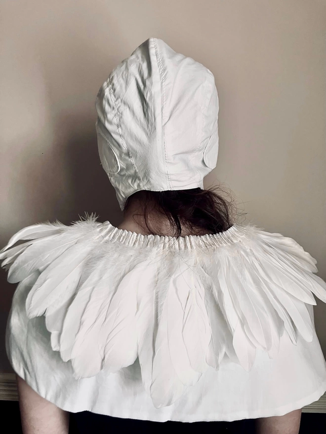 Image of the back of a girl facing a wall wearing a white feather cape and white flying hat