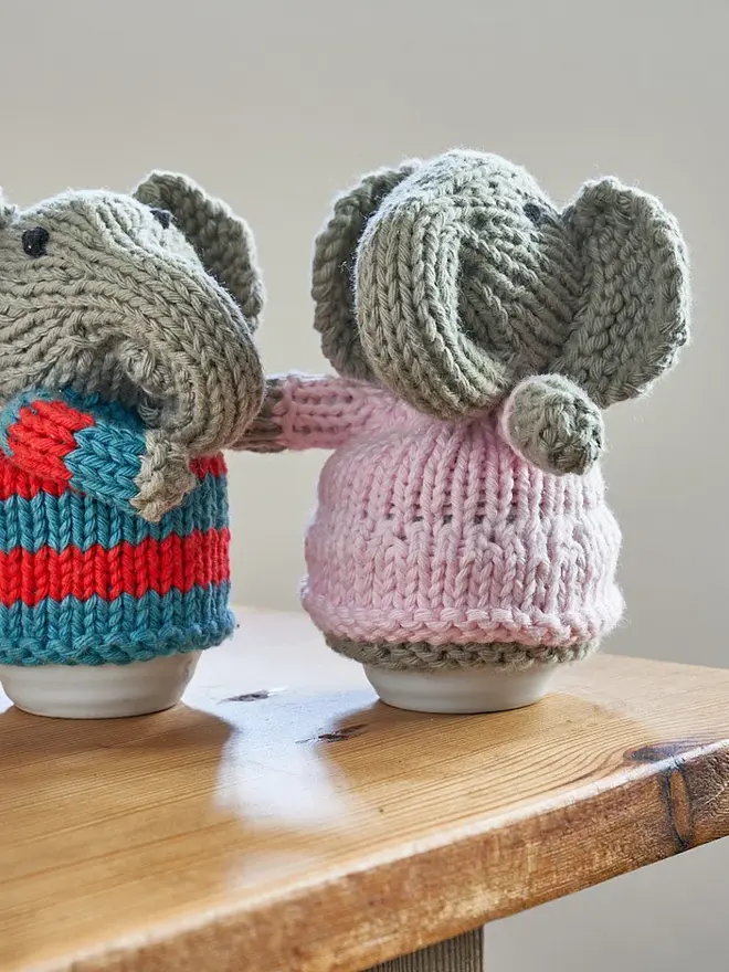 Egg Cosy Elephant Table Decoration (Set Of 2), a pair of colourful elephant cosies sitting on a wooden surface. 