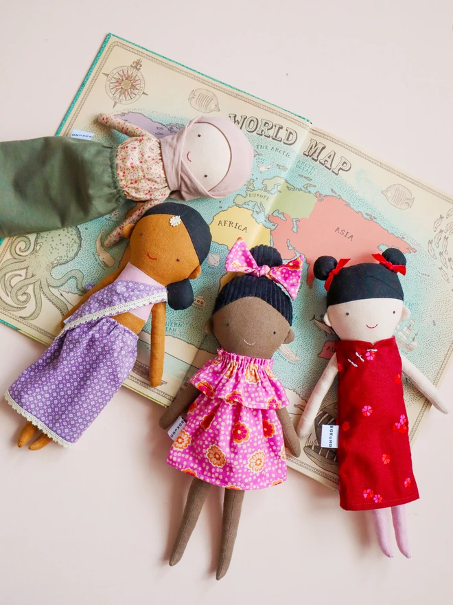Handmade fabric doll with medium brown skin tone, wearing a lilac patterned sari with lace trim and a pink top. The doll has black hair styled in a low side bun with a small white flower accessory on her forehead, simple stitched facial features. Next to Hijabi girl doll, african girl doll and chinese girl doll.