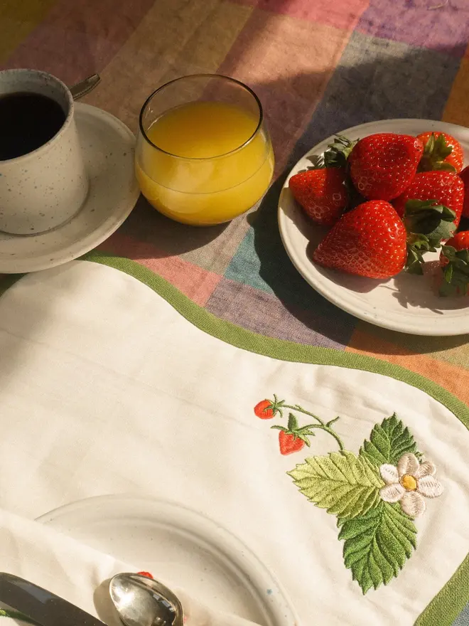 Embroidered strawberry table linen with green trim, styled for breakfast with fresh strawberries, coffee and orange juice.