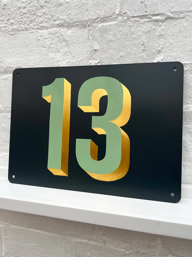 Sage green and gold leaf house number 13, on anthracite grey metal plaque, against a white brick wall.