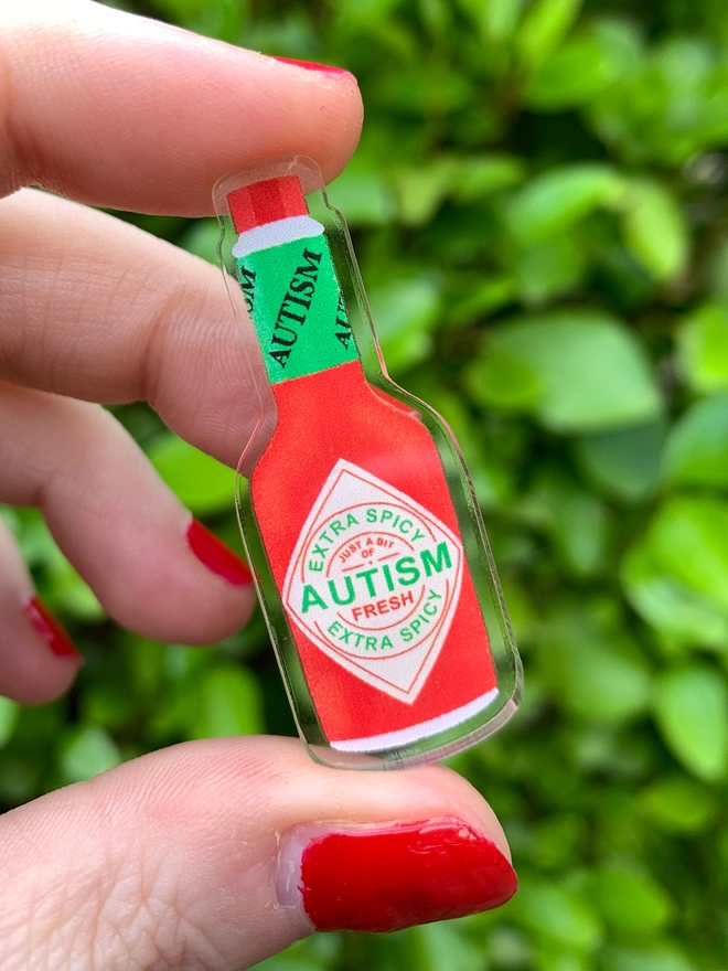 a hand holding an acrylic badge shaped and designed to look like a miniature hot sauce bottle. the bottle says 'fresh autism' on the label.