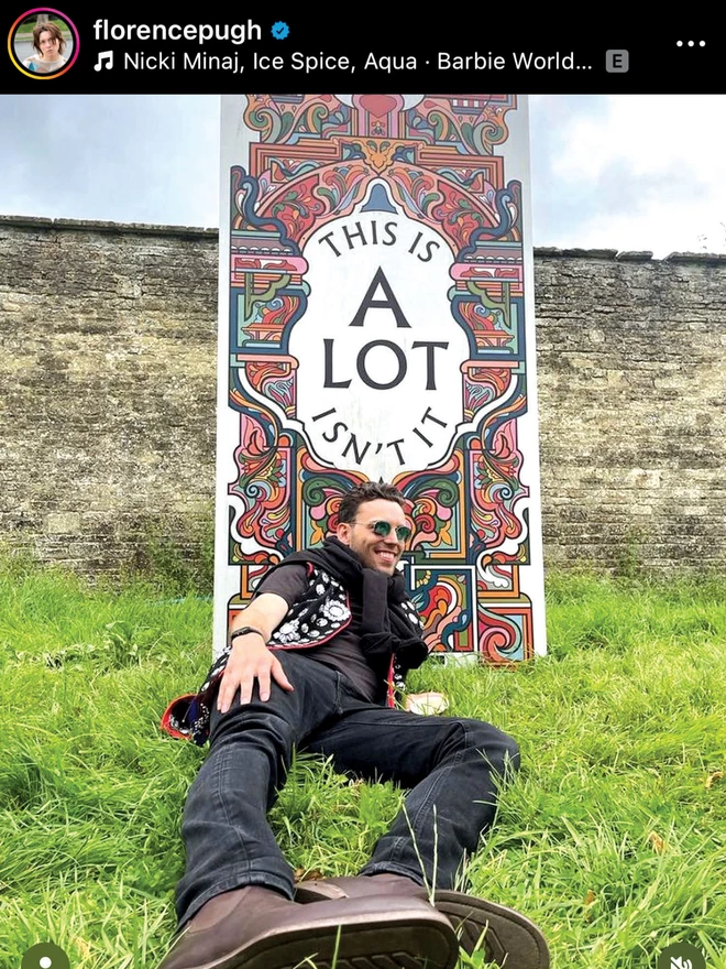 A screenshot of Florence Pugh’s instagram story of a white man in a black outfit laying on the grass in front of a huge banner print of the illustration, which rests against a stone wall. 