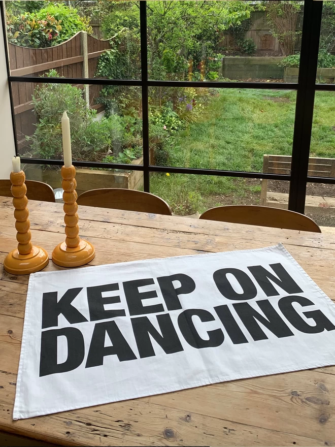 London Drying Keep On Dancing black screen printed text on white tea towel laying on wooden table with candlesticks behind it and view of garden through window panes in background