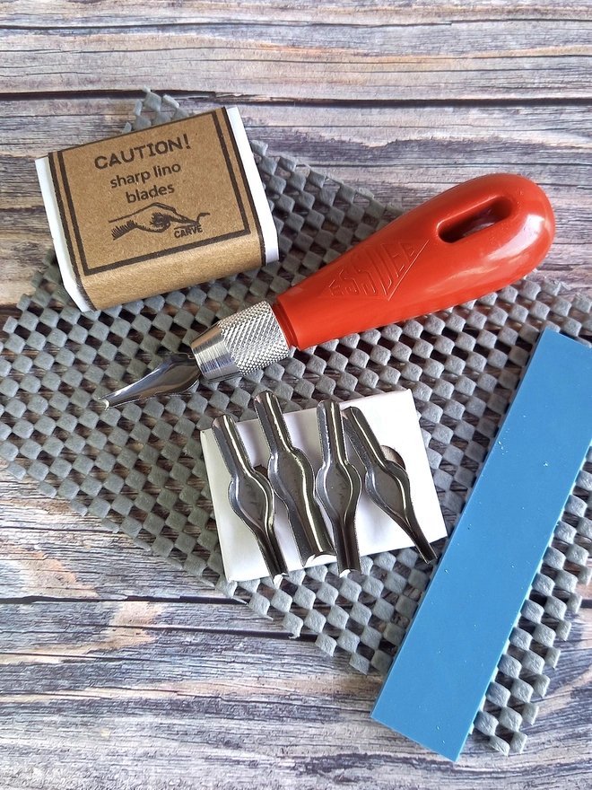 Lino Carving Printing Tool Kit In Presentation Box, carving utensils and strip of soft blue lino unpacked on a wooden table. 