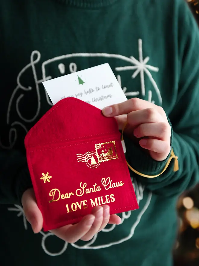 A child wearing a deep green jumper is holding a red felt personalised Christmas envelope that reads "Dear Santa Claus, Love Miles," with a festive letter card inside.