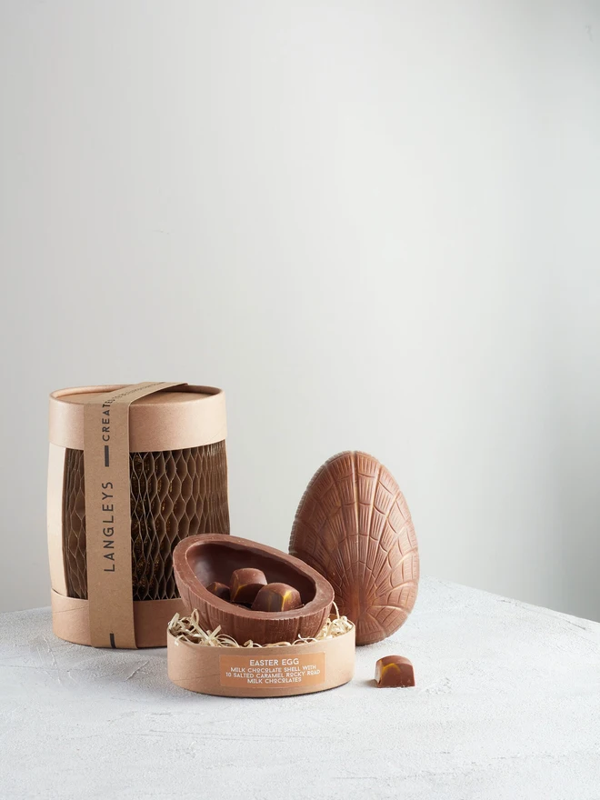 Large Milk Chocolate Easter Egg With Rocky Roads Chocolates, a chocolate egg sitting on a white surface against a plain backdrop. It is accompanied by some of the packaging, as well as another egg opened up to reveal the eggs within. 