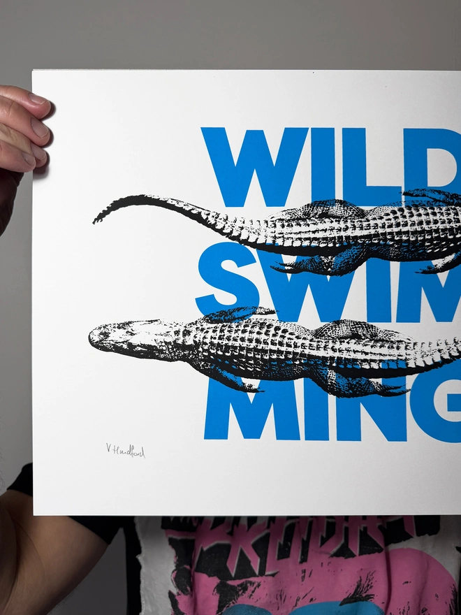 A humorous limited edition A3 screen printed poster. Featuring black alligators over blue 'wild swimming' typography. Showing close up of detail left edge.