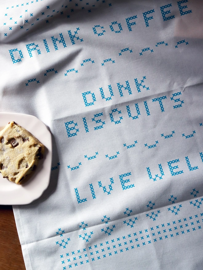 A Mr.PS Drink Coffee, Dunk Biscuits, Live Well cross-stitch typeface tea towel laid on a table with a shortbread cookie on a pink plate. 