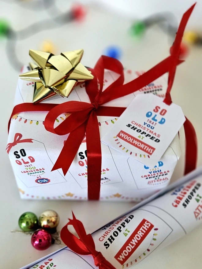 A rectangular present gift wrapped in a design that says so old that you Christmas shopped at Woolworths, C&A, Sock Shop, Tie Rack, Athena on an illustrated white tag with a red bow. The text is written in brightly colours with a string of coloured fairy lights.