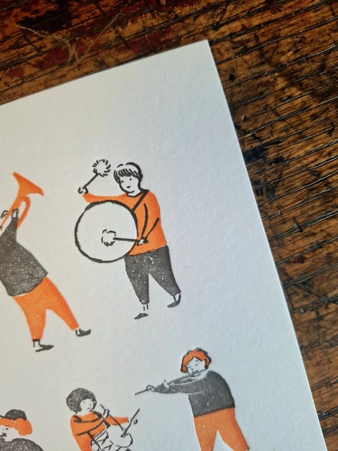 A close up of an illustration of a printed orange and black figure playing a drum, on a card lying flat on a wooden surface.