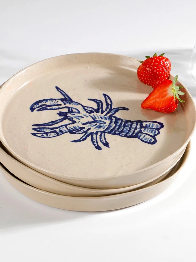 Handmade Ceramic Side Plate With Blue Lobster Design, a side plate with a blue lobster design sitting on a white table surface accompanied by a raspberry. 