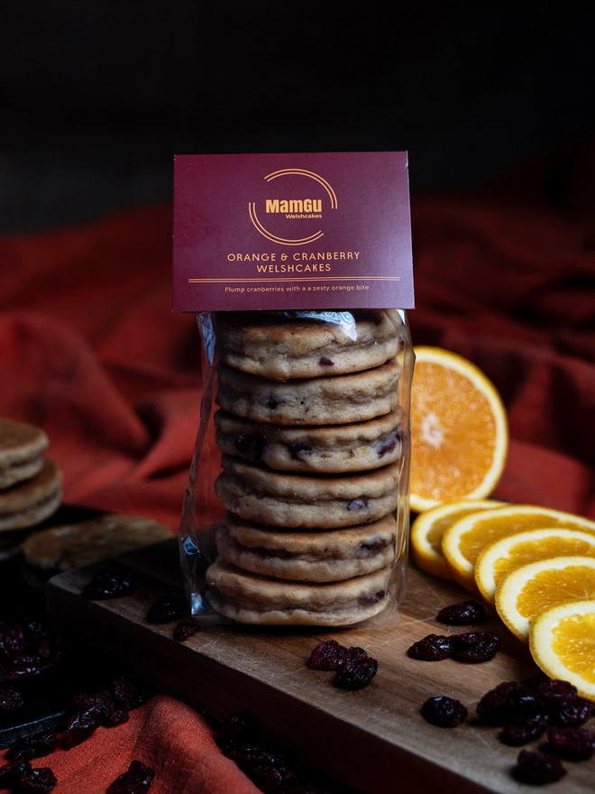 Packaged Orange and cranberry Welsh Cakes on a wooden chopping board with loose cranberries and sliced orange on an orange table cloth