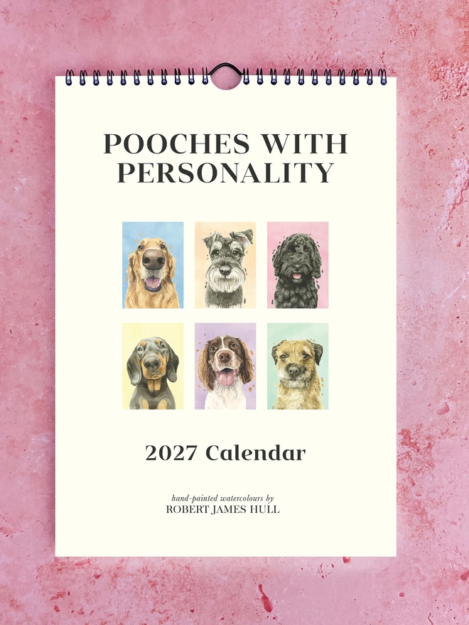2027 Pooches with Personality Calendar by Robert James Hull, laid down on a pink background, with the front cover visible.
