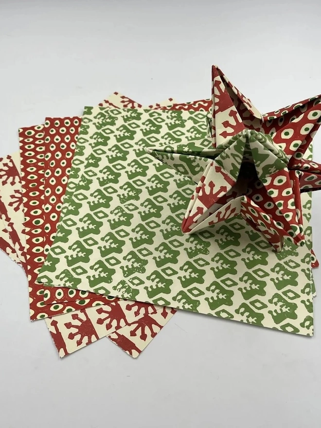Colourful Star Kit. A folded red and green paper star sitting on a fanned stack of matching printed paper squares. The festive craft kit papers feature cream backgrounds with red spot, red sunburst and green leaf block-print designs.