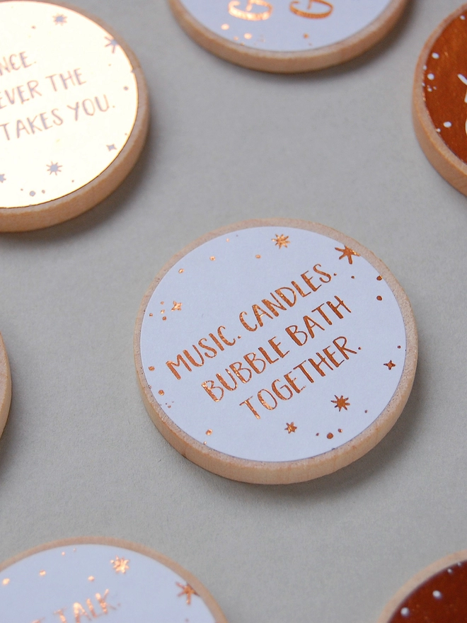 Several wooden tokens each with a copper foiled label that have a date night idea printed on lay on a grey background.