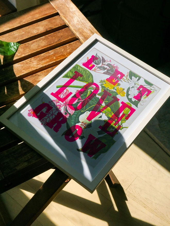 A framed limited edition artwork rests on a wooden surface, partially lit by sunlight. The letterpress print features a mix of bright pink and green leaves with the phrase "LET LOVE GROW" in bold, colorful letters rendered in fluorescent ink.
