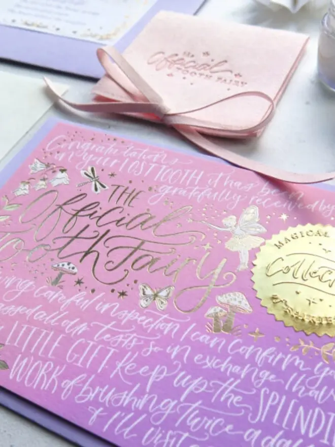 A pink-to-lilac ombre notecard with gold foil fairy, butterflies and toadstools and a "Collected" seal. The white calligraphy congratulates a child on their lost tooth, with a pink pouch behind.