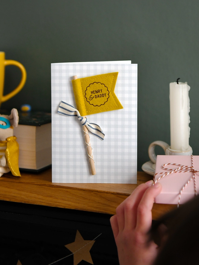 Personalised Father’s Day card with mustard felt flag reading “Henry & Daddy”, handmade keepsake card with ribbon and wooden stick detail. The card stands on a wooden mantle beside a book, mug, candle, and wrapped gift.