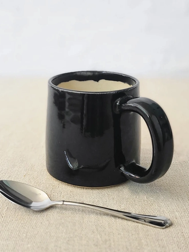 Ceramic Standard Mug, a colourful ceramic mug sitting on a plain surface against a grey backdrop. It is accompanied by a spoon. 