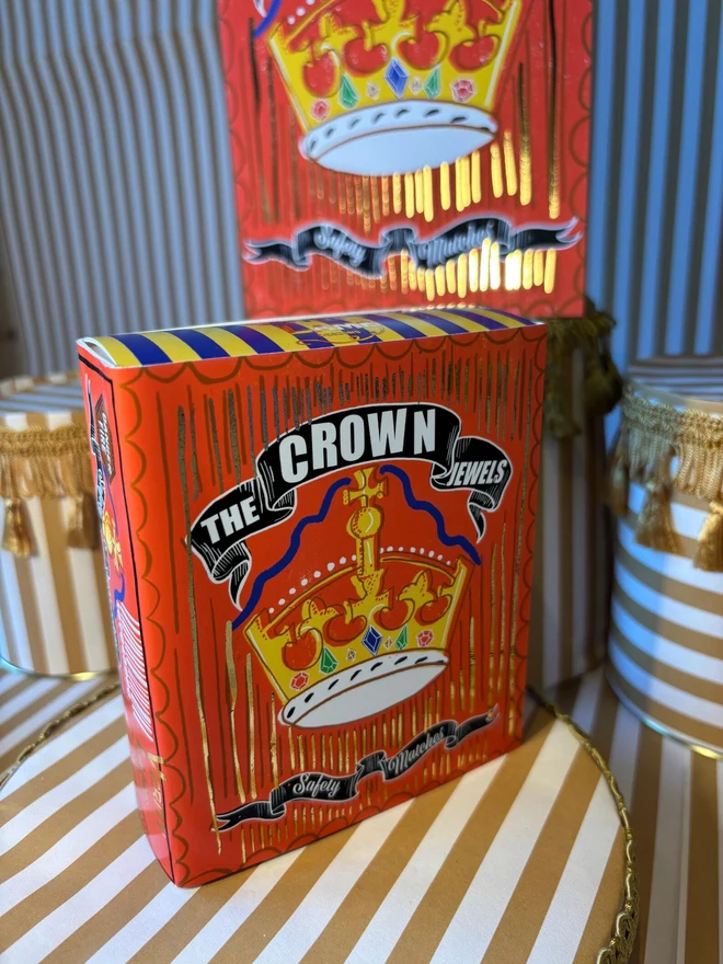 The Crown Jewels Matchbox, a red matchbox with a crown design sitting on a decorated surface accompanied by other accessories. 