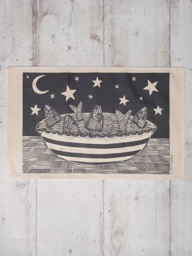 Picture of a tea towel with an image of a fish pie with the fishes gazing up at the stars, taken from an original lino print