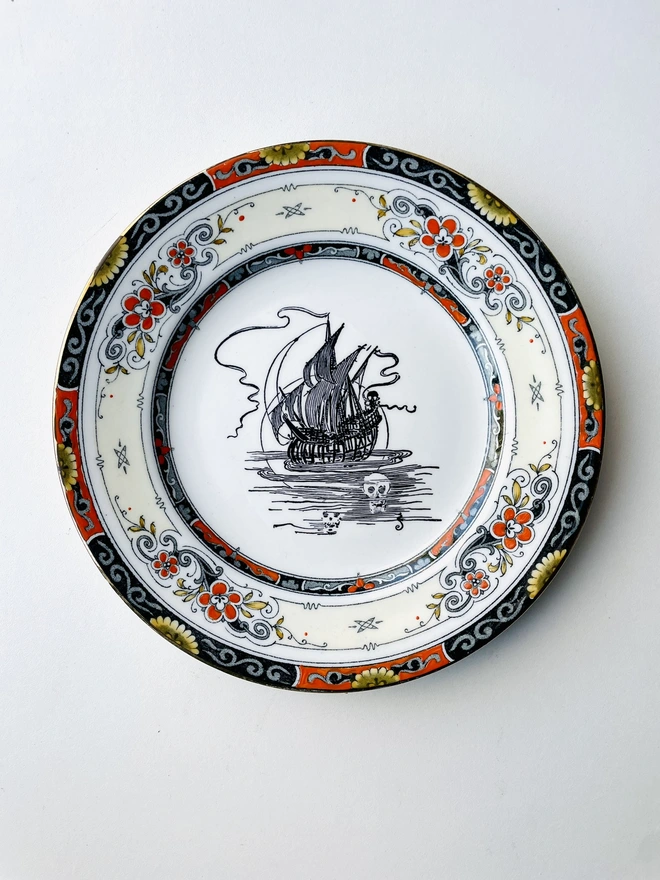 vintage plate with an ornate border of orange black and white pattern, in the centre of the plate is a vintage illustration of a ghostly ship at sea