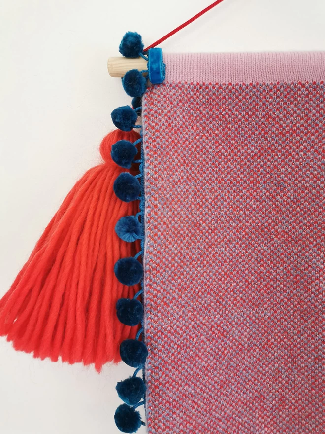 A product image showing the detail of the reverse of a scallop shaped wall hanging with teal pom pom trim edging. The back of the banner  is knitted into a red and blue birdseye check and an oversized red tassel can be seen hanging from one side of the wooden doweling.  The whole banner is suspended by a bright red nylon cord.