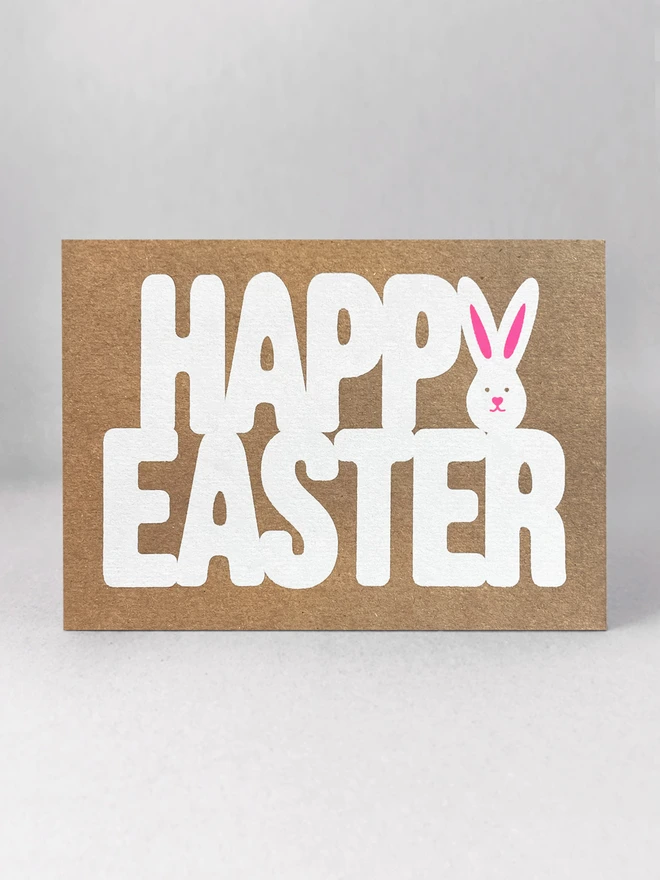 A landscape kraft card on a grey background. The words Happy Easter are screen printed in white ink. The letter Y of happy forms the shape of a white bunny. Neon pink ink is printed over the top to create the ears and the classic twitchy bunny nose.