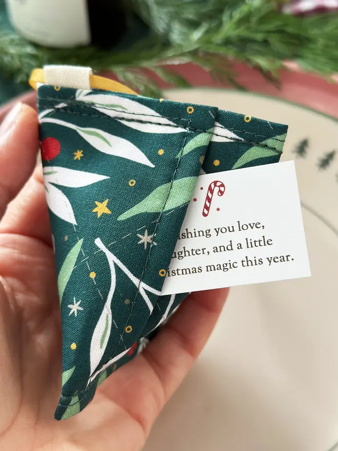 A handmade reusable fortune cookie Christmas decoration made from green festive foliage fabric is being held with a tiny Christmas wish card peeking out from the hidden pocket inside.