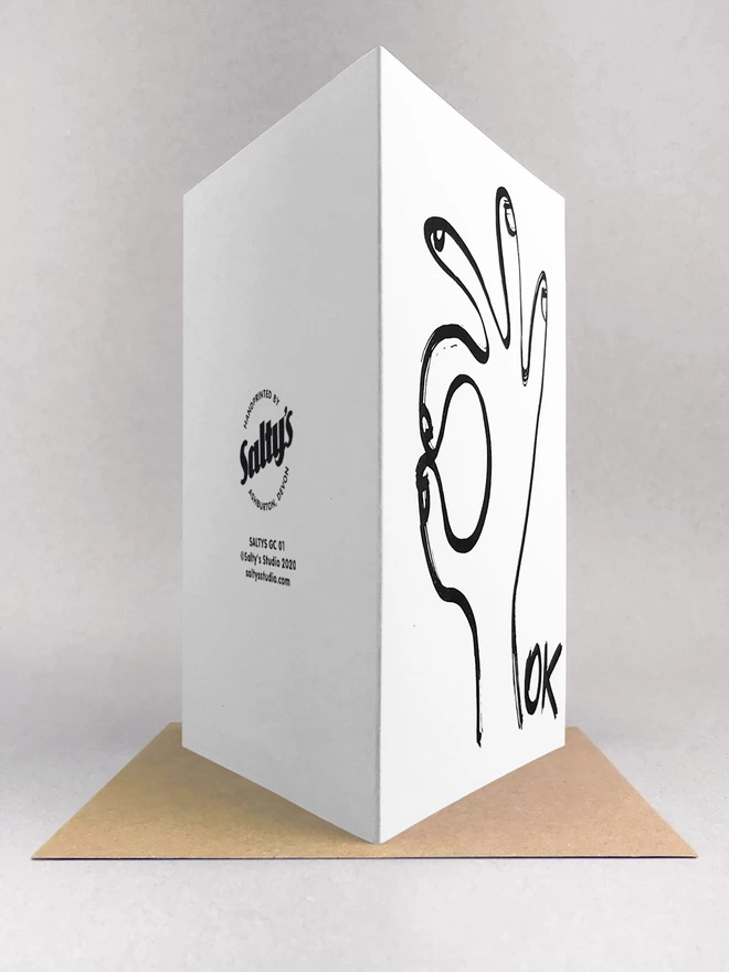 A white Ok Greetings Card with a black ink drawn hand and the word ok, rear view showing the logo on the back and stood on a Kraft brown envelope made by Salty's Studio.