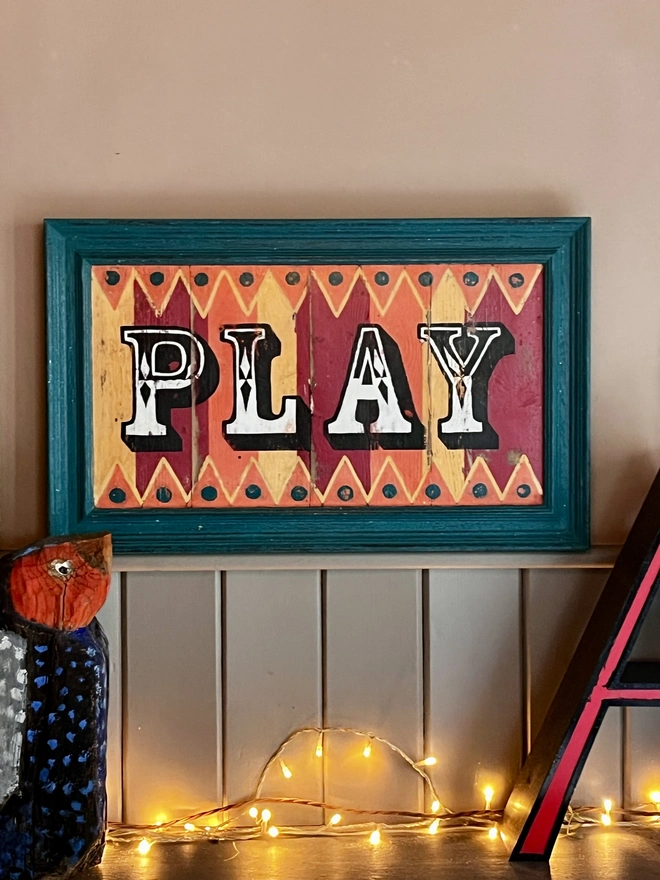 colourful play sign
made from reclaimed wood with a teal sign, hand painted fairground lettering and with orange pink and yellow stripes, with orange and yellow zig zag patterns  top and bottom
