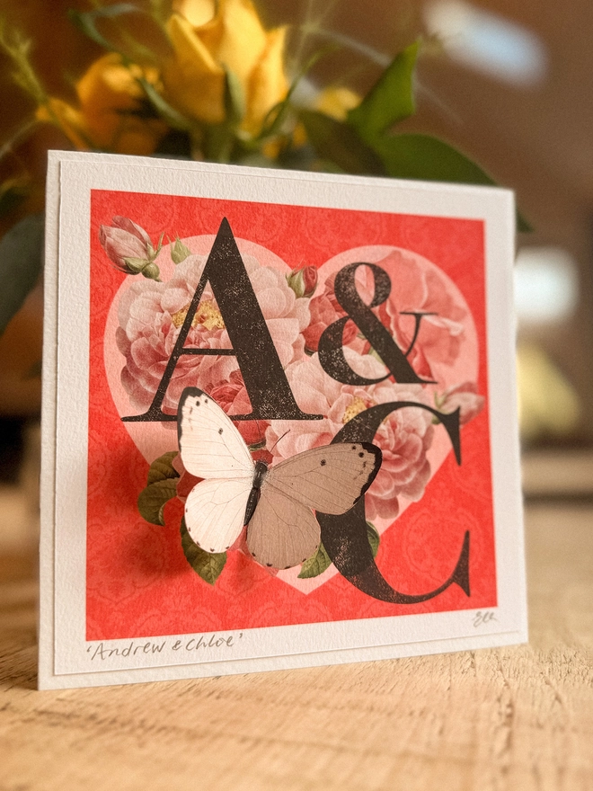 A handmade Ruby anniversary card featuring two initials of a couples names, place inside a heart design with vintage roses and on a red background. The recipients initials are hand written on the front of card and hand signed, A pink 3D paper cut butterfly sits on the heart and wings can be folded out for display.