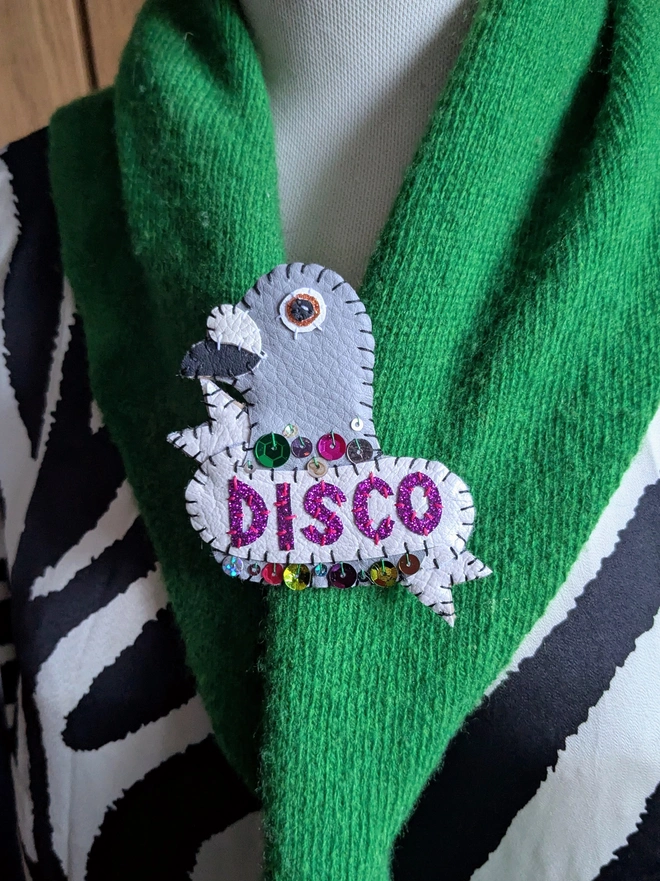 Hand-stitched white and grey faux leather pigeon brooch with purple glitter ‘DISCO’ lettering and colourful sequin details, pinned to a bright green scarf