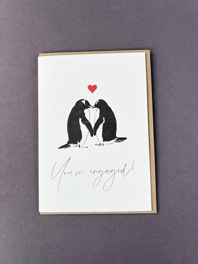 Two cute penguins touching beaks and holding flippers with a little red heart above them and "You're engaged" written underneath.